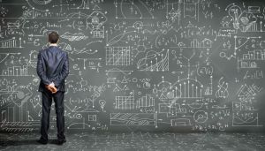15597956-Business-person-standing-against-the-blackboard-with-a-lot-of-data-written-on-it-Stock-Photo